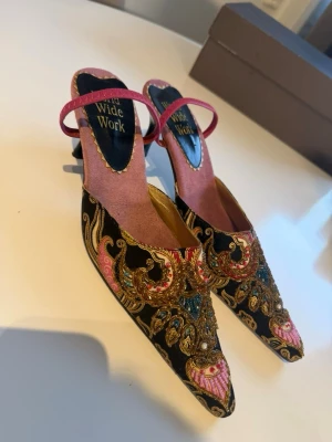 Brodderade pumps med pärlor och paljetter - Unika pumps från Wide Work med svart bas, broderade detaljer i guld, blått och rosa samt pärlor och paljetter. Skorna har spetsig tå, låg klack och rosa rem över foten. Perfekta för dig som vill sticka ut med färg och mönster.