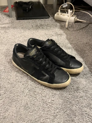 Svarta Golden Goose sneakers - Snygga svarta sneakers från Golden Goose med klassisk stjärndetalj på sidan. Skorna har svart läder och mocka, svarta skosnören och en vit, något smutsad sula för en cool vintage-look. Perfekta för dig som gillar streetstyle och exklusiva märken.