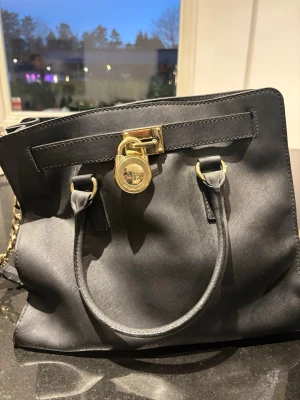 Svart handväska från Michael Kors - Snygg svart handväska från Michael Kors med guldfärgade detaljer och ett stort MK-lås framtill. Väskan har dubbla handtag och kedjerem på sidan. Tillverkad i ett tåligt material med elegant struktur. Perfekt accessoar för dig som gillar stilrena och lyxiga väskor.
