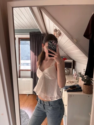 Vit spetslinne med rosett - Säljer ett vitt linne med spetsdetaljer och smala axelband. Toppen har en söt rosett framtill och ett luftigt, flowy tyg som ger en romantisk vibe. Perfekt till jeans eller kjol för en fräsch look i sommar.