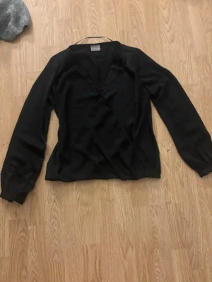 Svart omlottblus med långa ärmar - Stilren svart omlottblus med v-ringning och långa ärmar. Blusen har en enkel och elegant design som passar till många olika outfits. Tillverkad i ett lätt och mjukt tyg som ger en skön känsla. Perfekt för dig som gillar klassiska plagg med modern touch.