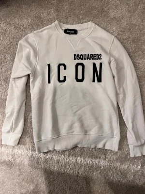 Vit sweatshirt från Dsquared2 ICON - Säljer en vit sweatshirt från Dsquared2 med svart text 'ICON' och logga på bröstet. Tröjan har rund halsringning, ribbade muddar och är tillverkad i mjukt material. Perfekt för dig som gillar streetwear och vill sticka ut.
