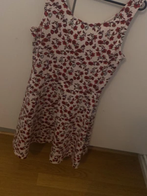 Vit blommig kortklänning - Söt vit kortklänning med röda och rosa blommor i ett tätt mönster. Klänningen är ärmlös och har en rundad halsringning. Materialet känns mjukt och stretchigt, troligen bomull eller polyester. Perfekt för varma dagar och ger en fräsch och ungdomlig look.