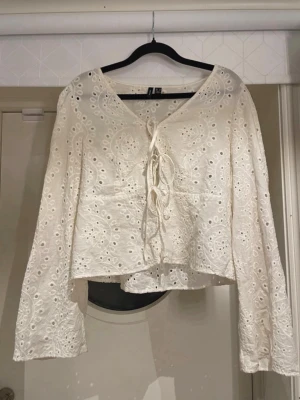Vit broderad blus med knytning - Superfin vit blus från veromoda med broderade hålmönster och v-ringning med knytning framtill. Blusen har långa ärmar och lätt utsvängd passform. Perfekt för en bohemisk look och tillverkad i bomull.
