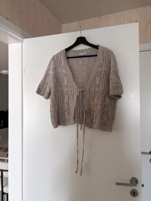 Beige stickad kortärmad kofta - Säljer en beige stickad kofta från House of Smith med korta ärmar och knytband framtill. Koftan har ett luftigt hålmönster och ribbade kanter. Perfekt att slänga över en topp för en avslappnad look.