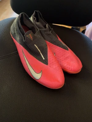 Nike Phantom fotbollsskor röd/svart - Säljer ett par Nike Phantom Vision fotbollsskor i rött och svart med stor vit Swoosh-logga på sidan. Dem är använda och lite sönder men annars det bara tape och dem funkar perfekt!