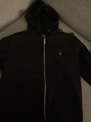 Svart hoodie från Polo Ralph Lauren - Svart hoodie från Polo Ralph Lauren med dragkedja framtill och klassisk röd logga broderad på bröstet. Huva med snörning och mjukt material som känns skönt mot huden. Perfekt för en chill och stilren look.