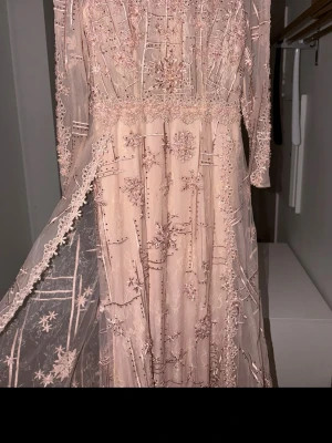 Ljusrosa långklänning med spetsdetaljer - Magisk ljusrosa långklänning med broderade spetsdetaljer och blommigt mönster. Klänningen har transparenta partier, lång ärm och vacker volangkant. Perfekt för dig som vill sticka ut med en romantisk look.