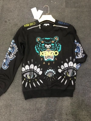Svart Kenzo sweatshirt med tigertryck - Cool svart sweatshirt från Kenzo med ikoniskt tigerhuvud och färgglada broderier framtill. Tröjan har detaljerade ögonmotiv och mönster på ärmarna samt dragkedjor vid axlarna. Kenzo-logga på axlarna. Perfekt statement-plagg för dig som gillar unika prints.
