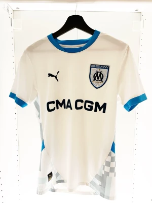 Vit Marseille fotbollströja Puma - Snygg vit fotbollströja från Puma med blå detaljer, Marseille-logga på bröstet och sponsortryck 'CMA CGM'. Tröjan har korta ärmar, rund hals och diskret rutmönster nertill. Perfekt för dig som älskar fotboll och vill sticka ut på planen.