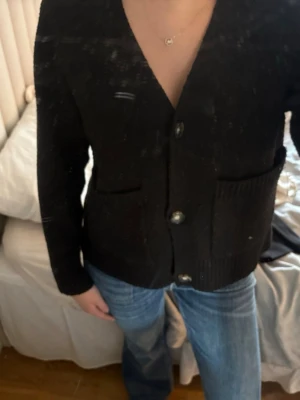 Svart kofta från Vero Moda - Svart kofta från Vero Moda med v-ringning och stora knappar framtill. Koftan har två fickor fram och är tillverkad i ett mjukt, stickat material. Perfekt att slänga på sig över en t-shirt eller linne för en chill look. 