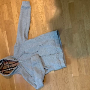 Grå hoodie från Burberry - Säljer en grå hoodie från Burberry med klassiskt rutigt foder i huvan och dragkedja framtill. Tröjan har två fickor och broderad logga på bröstet. Perfekt för en stilren och avslappnad look.