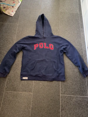 Blå hoodie från Polo Ralph Lauren - Mörkblå hoodie från Polo Ralph Lauren med stor röd POLO-text på bröstet. Klassisk känguruficka framtill och huva. Mjuk insida och avslappnad passform, perfekt för en chill stil. Kan gå ner i pris under snabb affär!