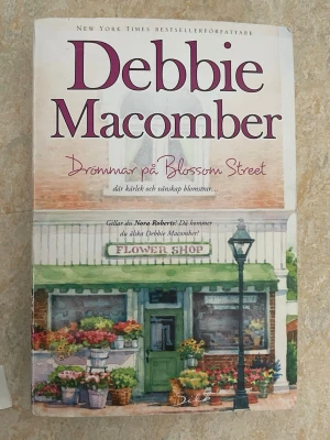 Drömmar på Blossom Street - En charmig roman av Debbie Macomber om kärlek och vänskap på Blossom Street. Omslaget har en akvarellillustration av en blomsterbutik med grönt tak och färgglada blommor. Perfekt för dig som gillar feelgood och varma berättelser.nypris butik kostar 280kr