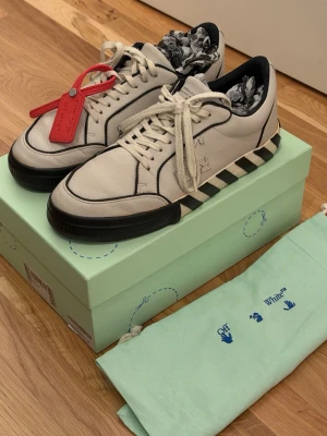 Off-White low top sneakers i läder - Off-White låga lädersneakers i off-white med svarta kontrastdetaljer och signaturrandig sula. Snörmodell med Off-White-detaljer på sidan, präglad Arrow-logga och ikonisk röd zip tie. Medföljer originalkartong och dustbag. Storlek 43. Mycket fint skick med lätta spår av användning. Kvitto finns dessutom!