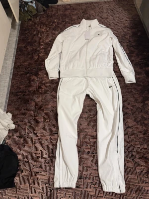 Vit träningsdress från Lacoste - Säljer en fräsch vit träningsdress från Lacoste med dragkedja framtill och diskret grönt krokodillogo på både jacka och byxa. Setet har smala gröna ränder längs ärmar och ben, ribbade muddar och hög krage. Perfekt för sport eller chill.