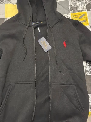 Svart hoodie från Polo Ralph Lauren - Svart hoodie med dragkedja från Polo Ralph Lauren. Klassisk design med röd broderad logga på bröstet, huva med dragsko och två fickor framtill. Tillverkad i mjukt bomullsmaterial som är skönt att ha på sig.