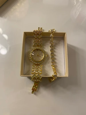 Guldig klocka med strass och armband - Säljer en riktigt blingig guldig klocka med matchande armband. Klockan har rund urtavla och hela bandet är täckt av glittrande stenar. Perfekt accessoar för dig som gillar att sticka ut och vill ha något extra. Kommer i ask.