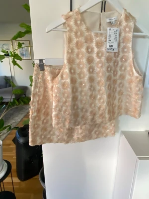 Set med topp och  kjol från H&M Edition - Säljer en beige kjol från H&M Edition med blommigt mönster och broderade detaljer. Kjolen har en kort passform och dragkedja i sidan. Linneblandning. Toppen är otroligt snygg, knäppe bak i nacken. 