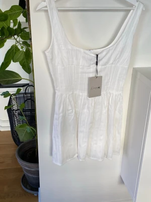 Vit kortklänning från Design By Si! Ny med prislapp  - Snygg vit kortklänning från Design by Si med subtilt randigt mönster och breda axelband. Klänningen har en feminin, lätt utställd form och är ärmlös, perfekt för varma dagar. Materialet känns lätt och luftigt, vilket gör den bekväm att bära. 