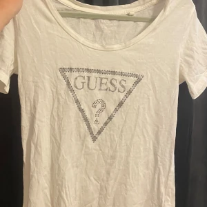Vit t-shirt från Guess med strass - Vit t-shirt från Guess med klassiskt triangellogo framtill dekorerad med strass. Rund halsringning och korta ärmar. Perfekt för dig som gillar stilrena plagg med lite extra bling. Den har en defekt vilket är ett pyttelitet hål som man inte tänker på. 