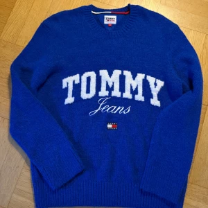 Blå stickad tröja Tommy Jeans - Snygg blå stickad tröja från Tommy Jeans med stort vitt TOMMY-tryck och broderad logga på bröstet. Tröjan har rund halsringning och ribbade muddar vid ärmslut och nederkant. Perfekt för dig som gillar klassisk streetwear med en sportig touch. Passar ca 175-180 cl