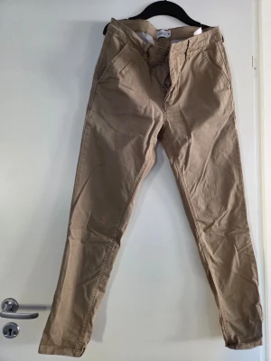 Beige chinos från Mouli - Snygga beige chinos från Mouli med klassisk passform och raka ben. Byxorna har fickor fram och bak samt bälteshällor. Materialet är mjukt och bekvämt, perfekt för dig som vill ha en stilren look.