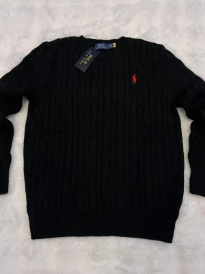 Polo Ralph Lauren Cable Knit Tröja - Svart kabelstickad tröja från Polo Ralph Lauren med klassisk rund hals och röd broderad logotyp på bröstet. Tröjan har ribbade muddar vid ärmslut och nederkant, perfekt för dig som gillar stilrena och tidlösa plagg.