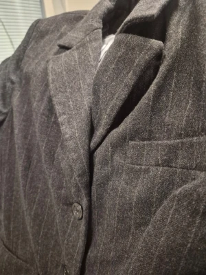 Mörkgrå randig lång kavaj med slag från Woman Collection 38 - Snygg mörkgrå kavaj med tunna vita kritstrecksränder och klassiska slag. Kavajen har två knappar framtill, två fickor med lock och en bröstficka. Perfekt för en stilren look och passar till skjorta eller t-shirt. Materialet ser ut att vara en mjukare ullblandning.