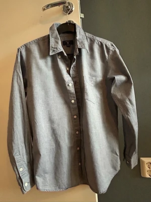 Blå skjorta från Gant med knappar - Säljer en klassisk blå skjorta från Gant med lång ärm och knappar hela vägen fram. Skjortan har en bröstficka med diskret logga och är tillverkad i mjuk bomull. 