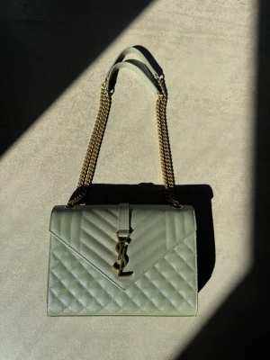 YSL Envelope Medium - ✨ Saint Laurent Envelope Medium i mintgrön – nyskick ✨  Säljer min exklusiva Saint Laurent Envelope Medium i den fina nyansen mintgrön (grain de poudre-läder) med guldfärgad YSL-logga och kedjerem. En tidlös och elegant modell som passar både till vardag och fest 🤍  Väskan är i nästintill nyskick och väldigt sparsamt använd – inga synliga slitningar.  👜 Modell: Envelope Medium 👜 Material: Grain de poudre-läder 👜 Färg: Ljusgrön / mint 👜 Mått: ca 24 x 18 x 6 cm Kommer tyvärr inte till användning.