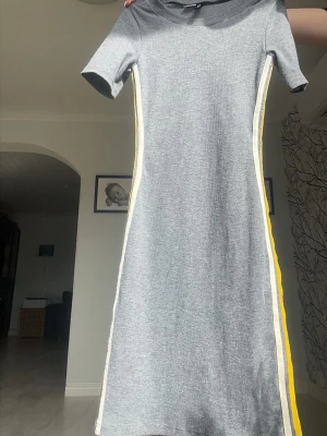 Grå kortärmad klänning med gula detaljer - Snygg grå kortklänning med korta ärmar och gula samt vita ränder längs sidorna. Klänningen är figur sydd och är tillverkad i ett mjukt, ribbat material som ger en skön känsla. Perfekt för dig som gillar en enkel men trendig stil🩶Kolla in min profil för fler fina och trendiga plagg!