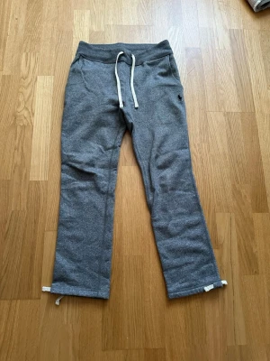 Grå mjukisbyxor från Polo Ralph Lauren - Grå mjukisbyxor från Polo Ralph Lauren med vit dragsko i midjan och raka ben. Byxorna har en liten broderad logga på höften och är gjorda i mjukt, fleeceliknande material. Perfekta för en chill och avslappnad stil.