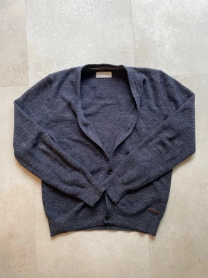 Mörkblå cardigan - Mörkblå cardigan med två knappar framtill och diskret mönster i tyget. Tröjan har v-ringad halsringning och ribbade muddar vid ärmslut och nederkant. Perfekt att slänga över en t-shirt för en avslappnad look.