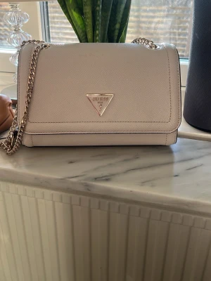 Beige handväska från Guess - Stilren beige handväska från Guess med guldfärgad kedjerem och klassisk trekantslogga framtill. Väskan är tillverkad i ett strukturerat konstläder och har en praktisk dragkedjeficka på baksidan. Har små defekter men inget man tänker på. Originalpris- 1600 kr. 