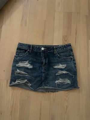 Blå jeanskjol med slitningar - Trendig blå jeanskjol med råa slitningar framtill och klassiska fickor. Kjolen har en kort passform och stängs med knapp och dragkedja. Perfekt för dig som gillar en avslappnad och edgy stil.