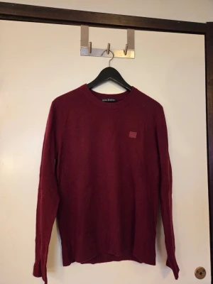 Vinröd stickad tröja Acne Studios - Vinröd stickad tröja från Acne Studios med rund hals och diskret logopatch på bröstet. Tröjan har långa ärmar och ribbade muddar vid hals, ärmslut och nederkant. Perfekt för dig som gillar stilrena plagg med premiumkänsla.