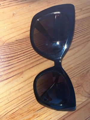 Svarta cat eye-solglasögon Dolce & Gabbana - Snygga svarta solglasögon från Dolce & Gabbana med cat eye-formade bågar och mörka glas. Sidorna pryds av DG-loggan i guld, vilket ger en lyxig touch. Perfekta för dig som vill sticka ut med en trendig accessoar.