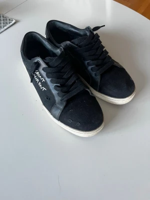 Svarta Saint Laurent sneakers - Svarta sneakers från Saint Laurent med vit sula och svart textilöverdel. Klassisk låg modell med snörning och handskriven logga på sidan. Perfekta för dig som gillar stilrena och trendiga skor med exklusiv känsla.