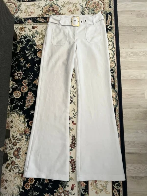 Vita bootcut byxor med bälte - Snygga vita bootcut byxor med breda ben och tillhörande bälte med fyrkantigt spänne. Byxorna har två framfickor och är tillverkade i ett kraftigare bomullstyg. Perfekta för en trendig och stilren look. Size M fits like XS.