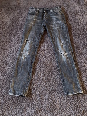 Grå slitna skinny jeans med hål - Säljer ett par grå skinny jeans med slitningar och hål på benen. Jeansen har fem fickor, dragkedja och knapp i midjan. Snyggt distressed look och smal passform, perfekta för en trendig streetstyle.