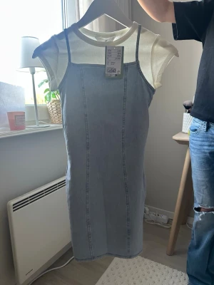 Ljusblå jeansklänning med ribbad topp - Snygg ljusblå jeansklänning med smala axelband och figurnära passform. Underklänningen är i ribbad, ljusvit bomull med korta ärmar. Perfekt för en trendig och avslappnad look.