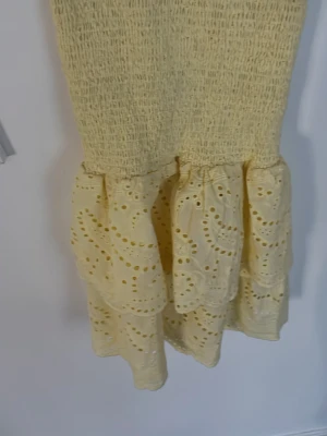 Gul volangklänning med broderi - Söt gul kortklänning med smockad överdel och dubbla volanger i kjolen. Kjolen har broderade detaljer och hålmönster som ger en romantisk känsla. Perfekt för varma dagar och sommarens alla tillfällen.