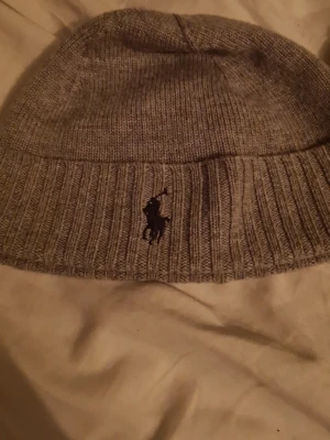 Grå stickad mössa från Polo Ralph Lauren - Snygg grå stickad mössa från Polo Ralph Lauren med klassisk broderad logga framtill. Mössan har bred ribbad kant och är tillverkad i mjukt material som håller dig varm. Perfekt accessoar för kyliga dagar.