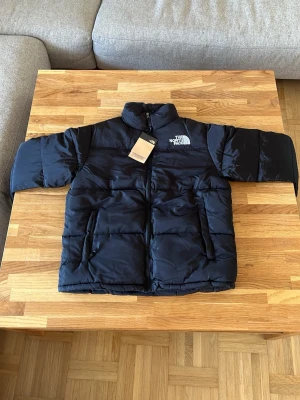Svart pufferjacka från The North Face - Säljer en svart pufferjacka från The North Face med hög krage och två stora framfickor med dragkedja. Jackan har en klassisk boxig passform och logga på både bröstet och ryggen. Perfekt för kalla dagar och riktigt snygg till streetwear-stilen.