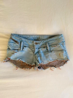 Ljusblåa jeansshorts - Supersnygga korta, lågmidjade, ljusblåa jeansshorts från Diesel med slitna kanter och låg midja. Shortsen har klassiska fickor, bälteshällor och dragkedja. Perfekta för varma dagar, semestern och en avslappnad stil.