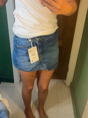 Blå jeanskjol  - Säljer en snygg blå jeanskjol med omlottdesign och knapp framtill. Kjolen är kort och har en klassisk denimlook som passar perfekt till vardags. Enkel att matcha med olika toppar för en trendig stil.