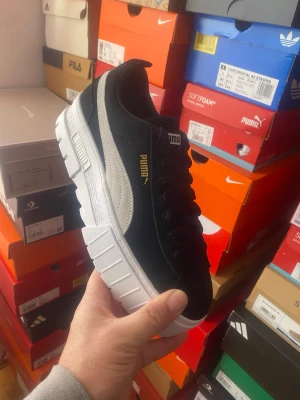 Svarta Puma sneakers med vit sula - Snygga svarta sneakers från Puma med kraftig vit sula och vit sidostrip. Skorna har klassisk låg profil, svarta skosnören och detaljer i guld på sidan. Perfekta för dig som gillar stilrena och sportiga skor.