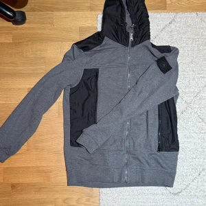Grå hoodie med svarta detaljer - Snygg grå hoodie med svarta detaljer och dragkedja framtill. Huva i svart material och svarta paneler på axlar och fickor ger en sportig look. Tillverkad i mjukt tyg med kontrasterande material på vissa partier. Perfekt för en avslappnad streetstyle.