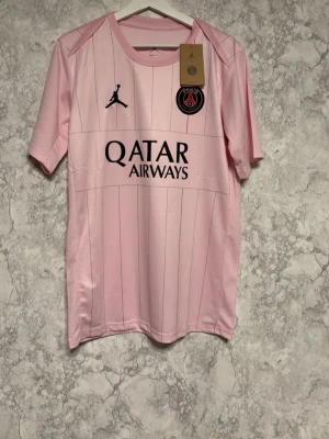 PSG rosa matchtröja Jordan M - Snygg rosa PSG matchtröja från Jordan med svarta detaljer och klubbmärke på bröstet. Tröjan har Dri-Fit material som andas och är perfekt för träning eller match. Qatar Airways-logga framtill och diskreta ränder ger en stilren look.